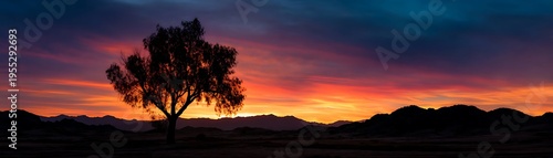 Dramatic fiery sunset illuminates the silhouette of a lone tree against dark mountain ranges