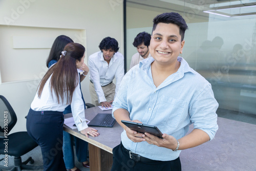 Smiling young indian male office employee holding digital tablet looking at camera while other colleagues working behind. Corporate job. Work culture.