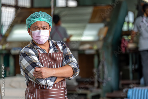 Confident textile factory worker posing with arms crossed in weaving machine mill. Portrait of male industrial staff wearing face mask and ear protection standing in fabric manufacturing plant.