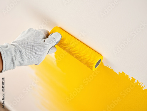 Hand in a grey work glove using a yellow textured paint roller to apply bright yellow paint onto a white wall surface