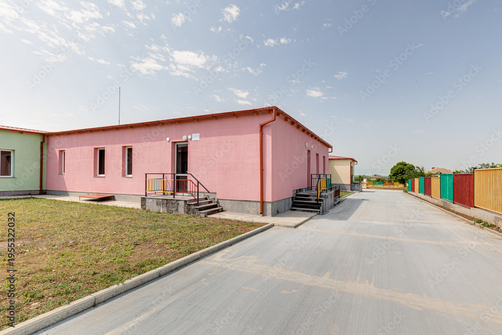 Fototapeta premium Outdoor view of a contemporary educational facility featuring bright colored railings, pink and beige buildings, concrete entrance stairs and a paved walkway under a clear sky.