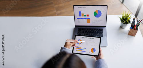 A bird's-eye view of a professional examining sales performance data on a tablet, while also considering a laptop report, highlighting the importance of analytics in business.