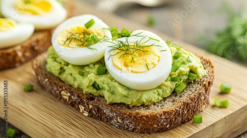 A close-up of a slice of whole grain bread topped with mashed avocado and halved boiled eggs, garnished with fresh herbs on a wooden board.