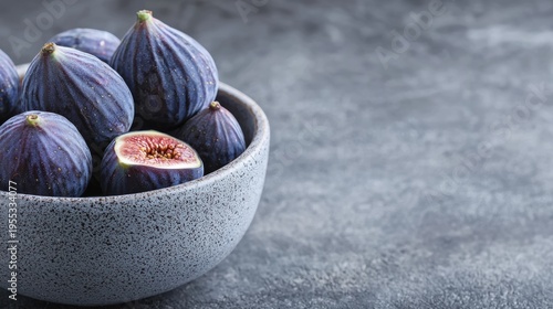 A bowl filled with fresh, ripe purple figs, one cut open to reveal the juicy red interior, set against a textured gray background.