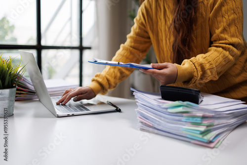 Fotografi A woman balances her laptop usage with organizing stacks of colorful paperwork, blending modern technology with traditional office tasks in a bright environment