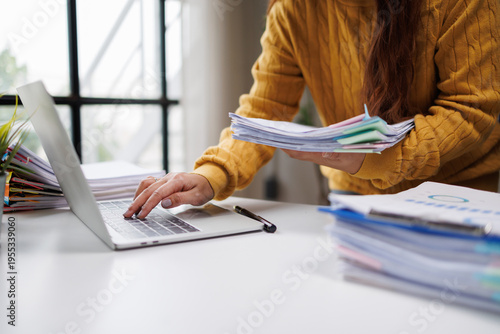 A woman in a yellow sweater organizes several stacks of documents while using her laptop, representing an engaging work atmosphere in a modern office setup.