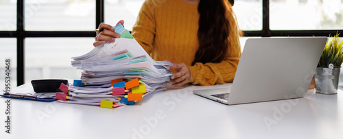 A woman efficiently organizing a tall stack of documents with colorful tabs while working on her laptop in a bright, modern office space filled with natural light.