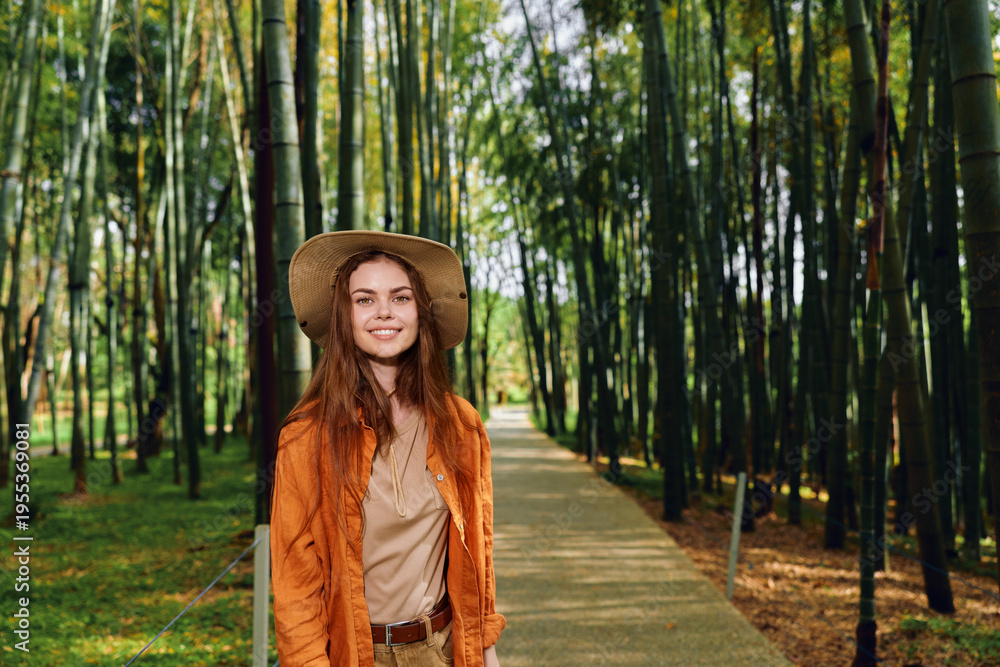 Fototapeta premium Woman smiling in straw hat and orange jacket walking on wooden path through bamboo forest, portrait of traveler in nature with sunlight filtering through tall bamboo stalks.