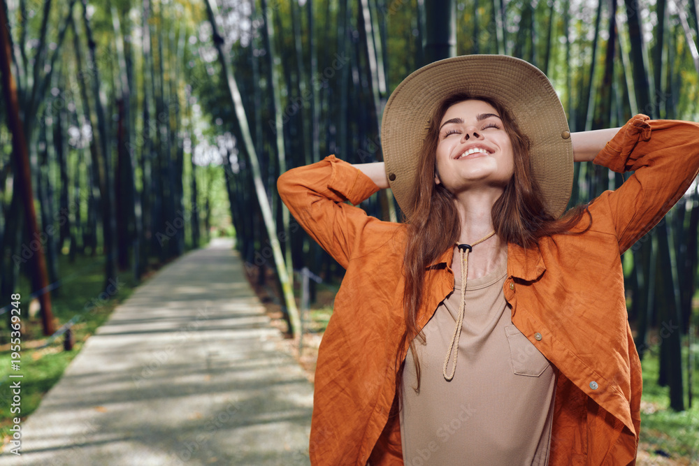 Fototapeta premium woman smile hat jacket park path trees nature enjoying sunlight and serenity, young woman in orange jacket with wide hat breathing fresh air on tree-lined path for relaxed outdoor portrait