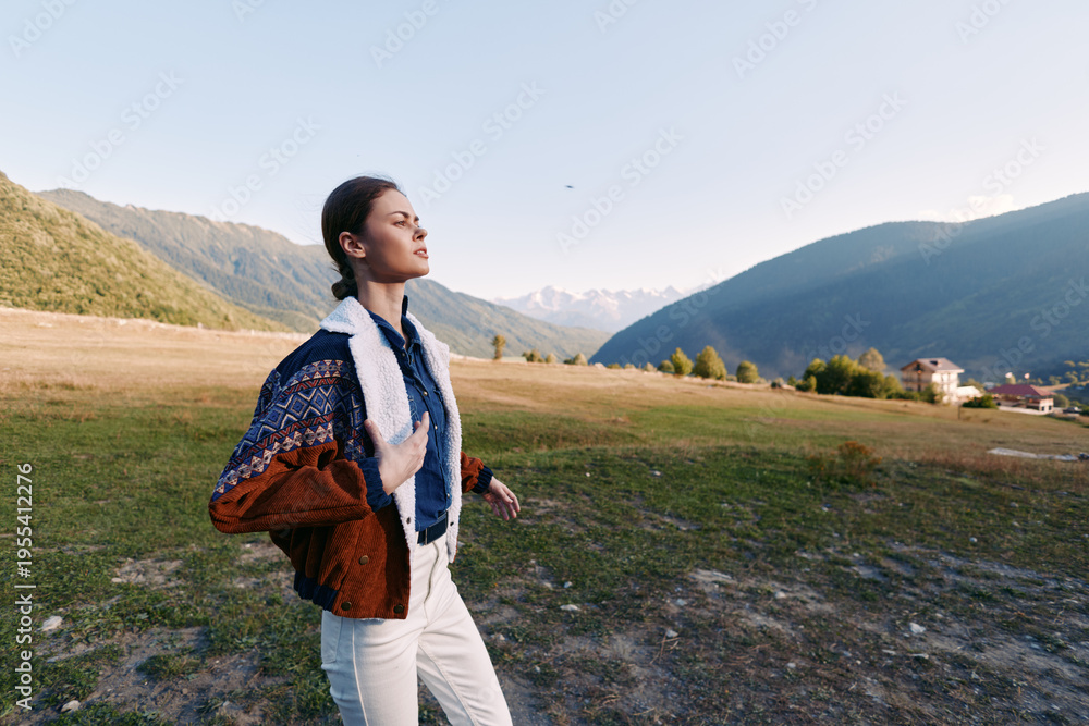 Fototapeta premium Woman in nature standing in an open field beneath distant mountains, eyes closed breathing deeply, casual portrait wearing a chunky sweater and scarf, peaceful relaxation outdoors.