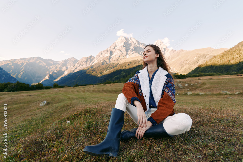 Obraz premium Woman sitting on meadow in front of mountain landscape, outdoor portrait of young woman wearing jacket and boots, relaxed pose in nature with scenic horizon and clear sky.