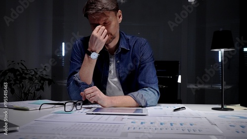 Schilderij op canvas Young businessman with glasses sits at a dark office desk late at night, overwhe