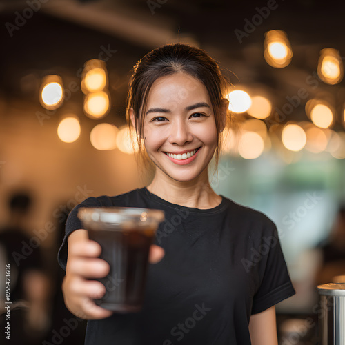 A beautiful Asian woman smiles, wearing a black shirt and holding a glass of iced coffee in a coffee shop.