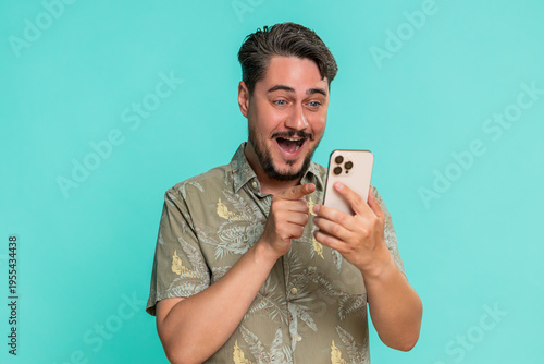 Middle-aged man joyfully laughing, looking at smartphone screen, reacting to funny memes or viral online videos. Young adult guy isolated on blue background enjoying digital humor and entertainment