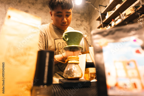 Young Asian man barista manual coffee brewing pour over method specialty coffee shop warm ambient light focus concentration cafe counter coffee equipment lifestyle scene cozy atmosphere