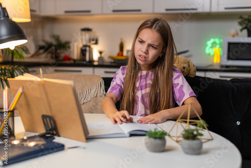 Teenager girl at night home reads problem from book for homework feels confused holds head sighs. Caucasian child kid sitting at table at evening puts notebook aside looks tries to understand slowly.