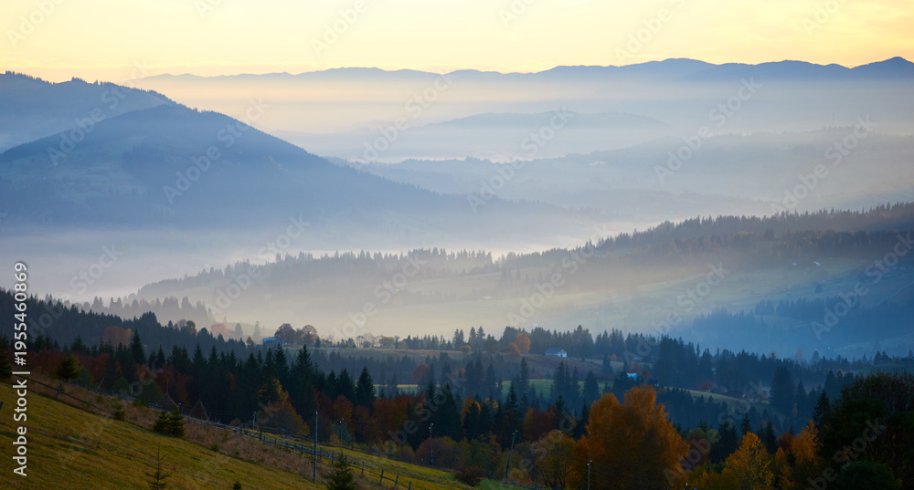 Obraz premium Morning sunlight streams through mist, illuminating serene mountain village nestled among forested hills. Light creates ethereal rays that gently touch autumn foliage and houses scattered on slopes.