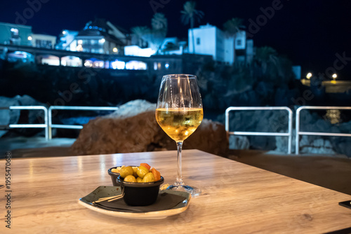 Glass of white wine from Tenerife served in cafe by ocean in Puerto de la Cruz at night with view on old houses, evening dining, winter vacation in Spain