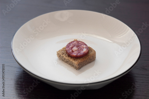 A small sandwich with a slice of sausage on a slice of rye bread in the center of a white plate. Minimalistic presentation and an ironic contrast. Very simple.