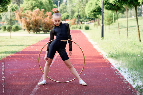 Young gymnast practicing rhythmic moves using a hula hoop on an outdoor track surrounded by greenery. Young Gymnast Practicing Rhythmic Routine Outdoors With Hula Hoop on Track