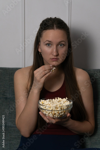 Sitting comfortably on a grey sofa, a young woman savors a bowl of popcorn while engrossed in a captivating movie, her expression reflecting the suspense and excitement of the film. Vertical photo