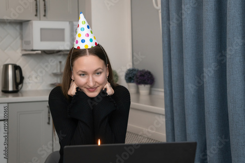 Young woman celebrating a birthday online, wearing a colorful party hat and smiling while using a laptop. A lit candle sits in front of the screen as she makes a wish during the virtual event
