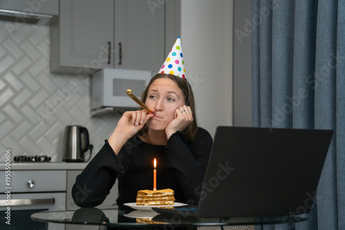 Sad woman wearing party hat blowing party horn and celebrating birthday online using laptop at home in kitchen, with piece of cake and candle