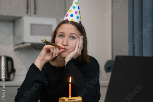 Young woman wearing party hat blowing in blowing a festive horn and looking bored while celebrating birthday online using laptop at home during lockdown