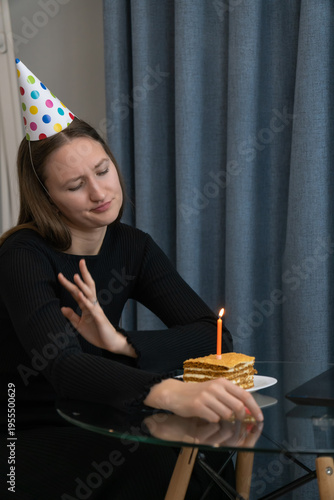 Sad young woman wearing party hat celebrating birthday online during lockdown looking at her laptop and holding a slice of birthday cake with a burning candle. Vertical photo