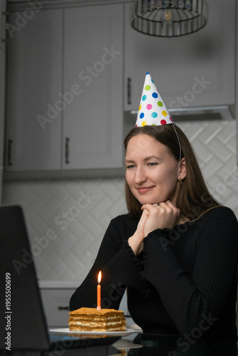 Young woman celebrating her birthday online, wearing a festive party hat while making a wish with a lit candle on a delicious cake, all set up in front of her laptop at home