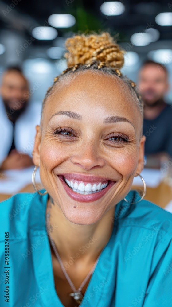 Obraz premium A confident healthcare professional woman in teal scrubs smiles warmly. She is indoors in an office setting with blurred colleagues in the background, appearing to be in a meeting during the day