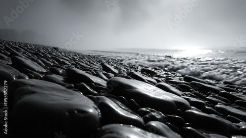 Black and white seaside landscape with shining sun over waves lapping pebble beach on a misty day