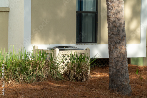 Obraz na płótnie Outdoor central air conditioning unit behind a decorative lattice fence, surrounded by green foliage and pine needles, next to a building wall and a tropical palm tree trunk
