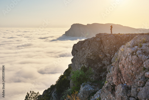 Schilderij op canvas Landscape of High Rock Cliff Above Sea of Clouds at Golden Hour Sunset with Girl