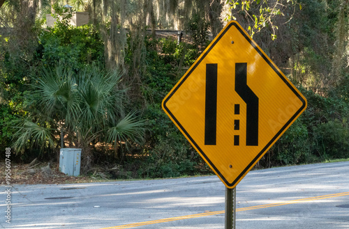 фотографія Bright yellow diamond-shaped road sign warning drivers of an upcoming lane merge or road narrowing on a scenic route with lush green foliage and Spanish moss