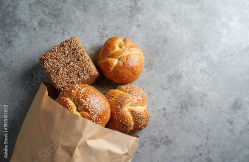 Assortment of fresh baked goods spilling from a brown paper bag onto gray textured surface. Includes seeded bread loaf, sesame buns, and plain rolls, offering delicious breakfast or snack options.