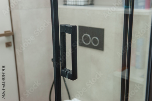 Glass shower door with black frame and minimalist handle in modern bathroom. Stylish interior design detail with functional wall mounted flush button. Contemporary home renovation concept.