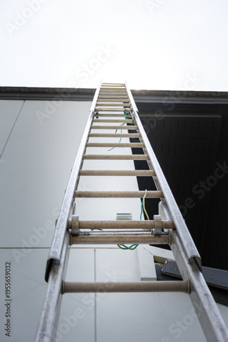 An aluminium ladder is leaning on the house roof, using for climbing ascent up to roof top for building activity susch as installing the solar cell. Close-up and selective focus at ladder's part.