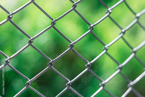 Metal mesh delimiting a private property. Close-up of a chain-link fence with a blurred green background. The texture of the metal is visible, creating a contrast with the soft foliage behind it.