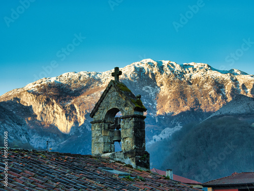 Rural Bell Gable against Snowy Mountain Peak - Rustic stone bell gable and cross on a tiled roof, sharply contrasted with a vast, sunlit, snow-capped mountain backdrop.