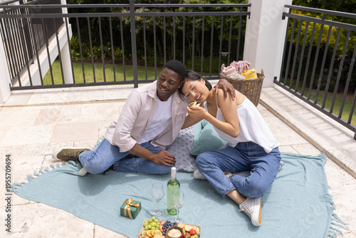 Diverse couple sitting on light blue blanket on rooftop patio sharing fruit and wine