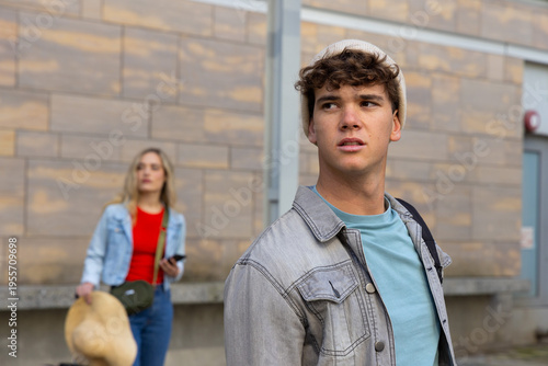 Teenage friends standing on urban street in light denim jackets, holding smartphone and straw hat