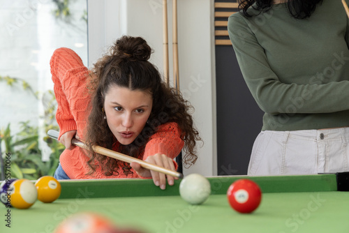Female friends playing pool in game room, one wearing orange sweater aiming cue stick
