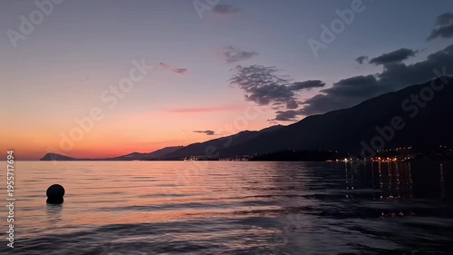 Serene Coastal Sunset Over Calm Sea with Silhouetted Mountains and Distant Town Lights Reflected