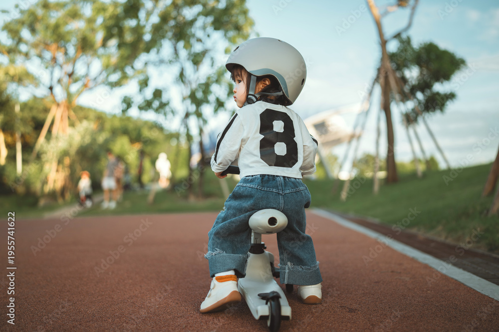 Fototapeta premium Rear view - cute Asian toddler girl wearing a safety helmet while learning to ride a balance bike at a public park during a sunny afternoon, representing early childhood development, outdoor