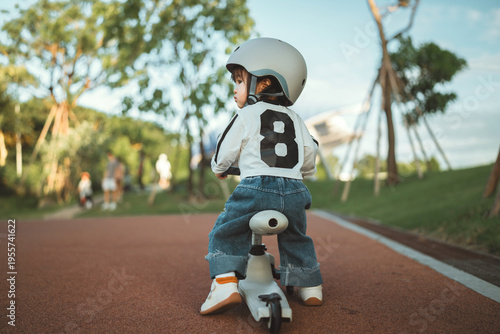 Rear view - cute Asian toddler girl wearing a safety helmet while learning to ride a balance bike at a public park during a sunny afternoon, representing early childhood development, outdoor
