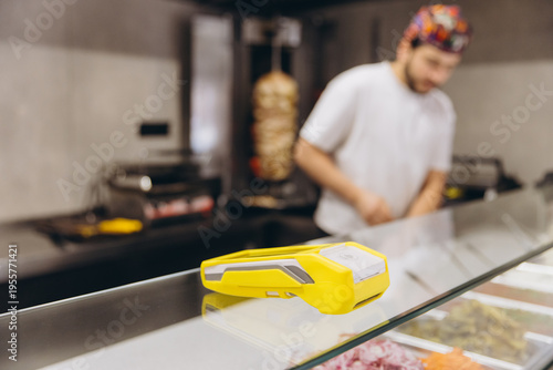 Yellow POS terminal on a glass display counter in a fast food restaurant, with a worker and a vertical rotisserie
