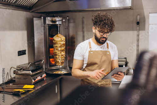 Arab man chef managing online orders on tablet in fast food restaurant kitchen with shawarma cooking