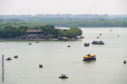 A panoramic view of tour boats navigating the calm waters of Kunming Lake near a scenic island at the Summer Palace in Beijing, with a hazy city skyline in the background.