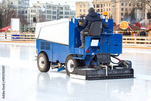 Zamboni cleans ice rink in city park during winter day with festive lights in background